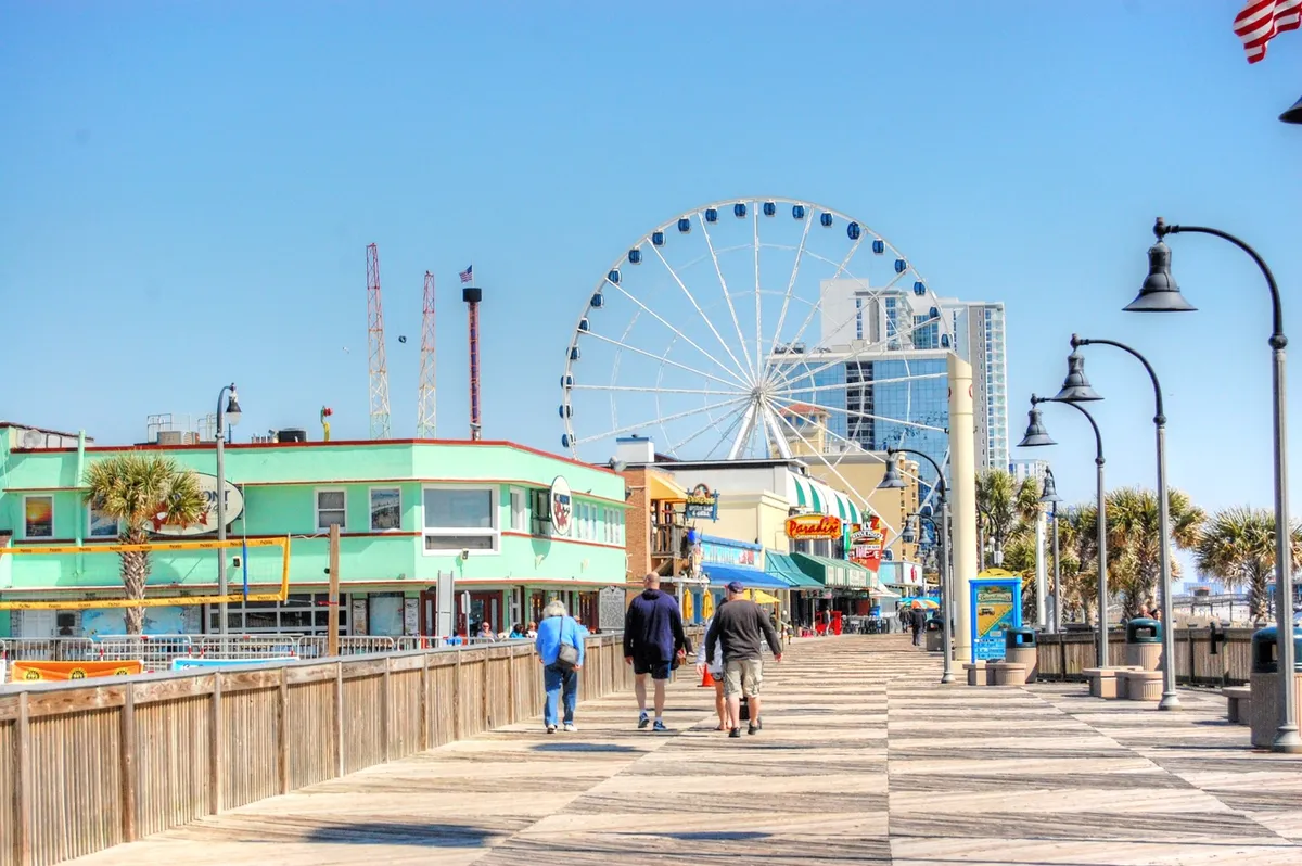 A joyful exterior view of a lively boardwalk featuring a Ferris wheel, colorful storefronts, palm trees, and people strolling—capturing Myrtle Beach's classic entertainment atmosphere.