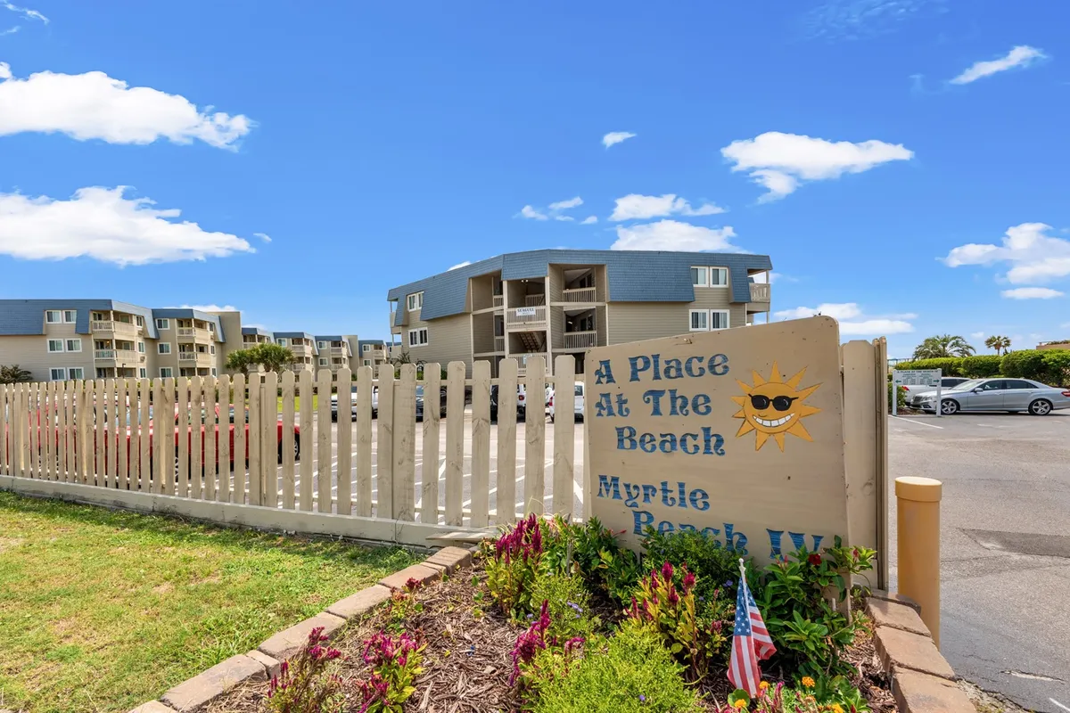 A cheerful exterior shot featuring a sign with a sun graphic, lush landscaped garden, and buildings in the background. The scene evokes the vacation vibe of Myrtle Beach, with the promise of beach access and outdoor fun just moments away.