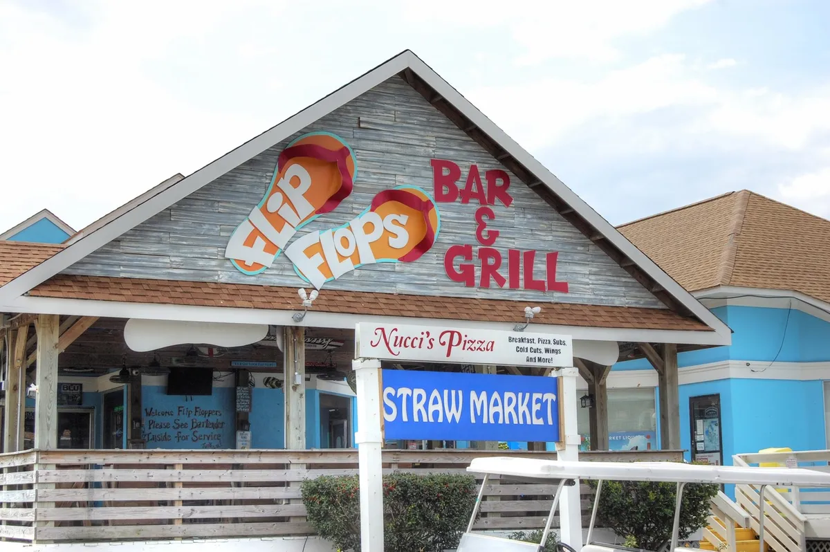 A lively streetscape featuring signs for Flip Flops Bar & Grill, Nucci's Pizza, and the Straw Market, set against a sunny backdrop. The scene suggests a welcoming area for casual dining, outdoor seating, and browsing local shops, with a relaxed coastal vibe.