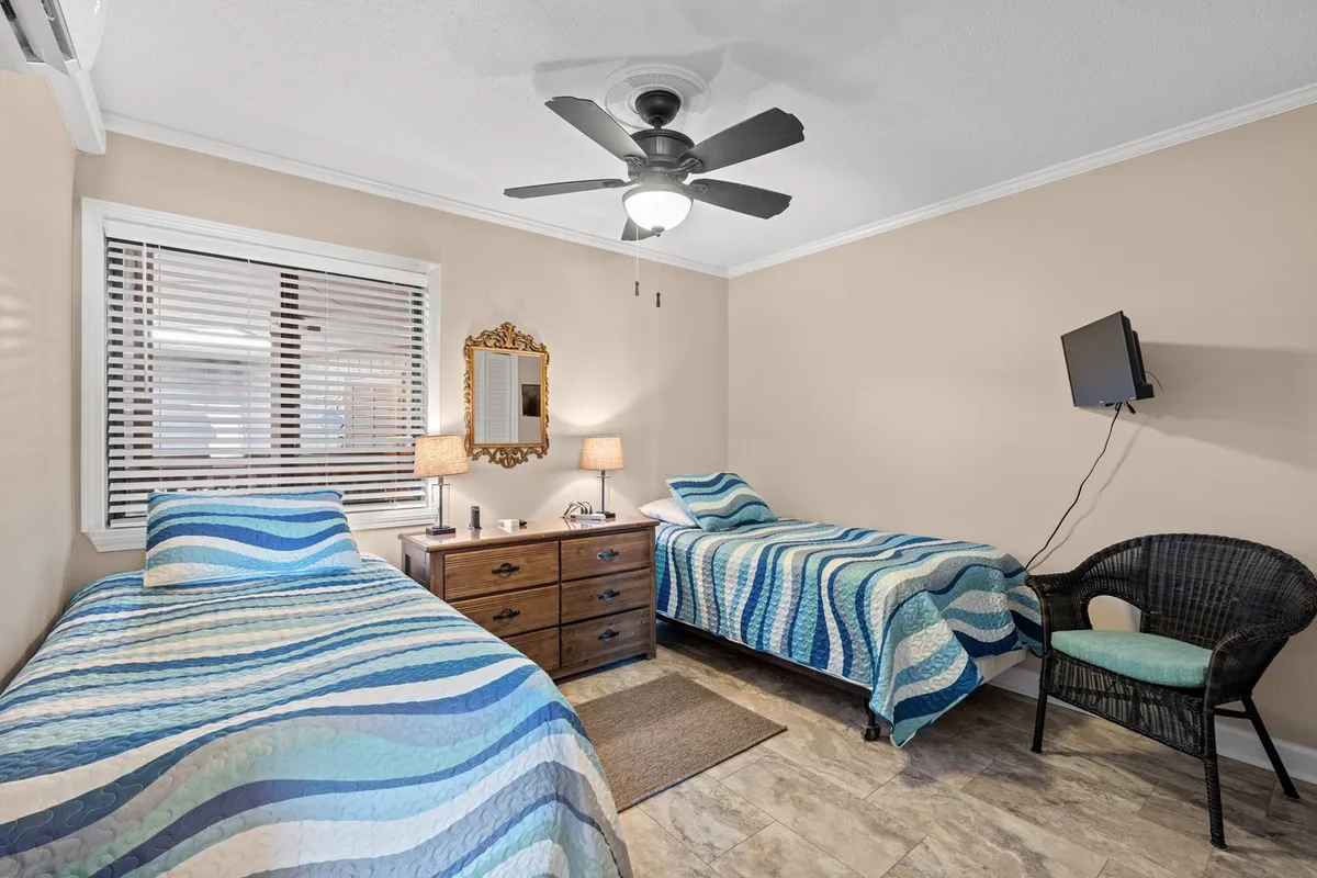 A cozy second bedroom with two twin beds covered in patterned quilts, a wooden dresser with an ornate mirror, table lamps, and a wicker chair. The layout is perfect for kids or friends, fostering both comfort and a sense of independence.