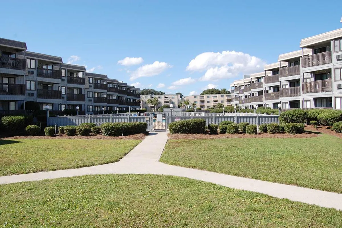 Exterior view of the building's pool area, lush landscaped gardens, and multiple balconies visible on the apartment units. The scene is sunlit and relaxed, with a communal but tranquil feel.