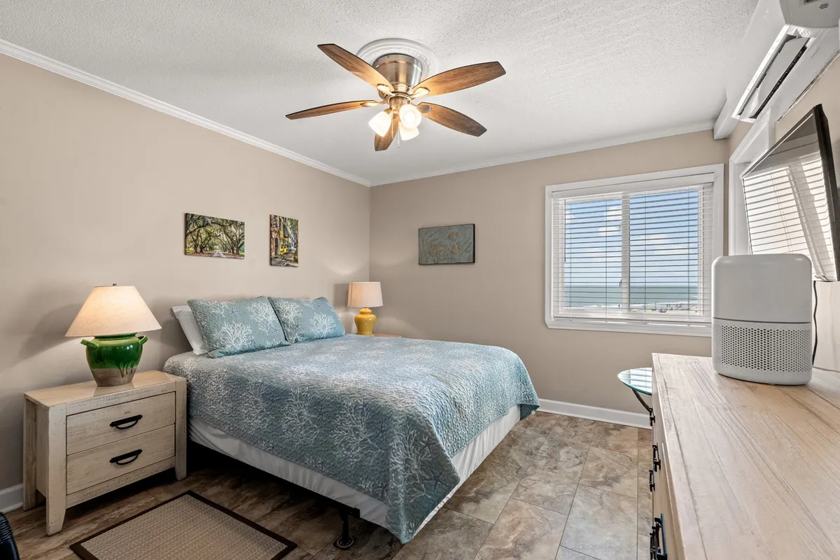 A serene bedroom scene featuring a neatly made bed, a window letting in natural light, decorative artwork above the headboard, and a bedside lamp. The space feels calm and uncluttered, ideal for relaxation and restful sleep.