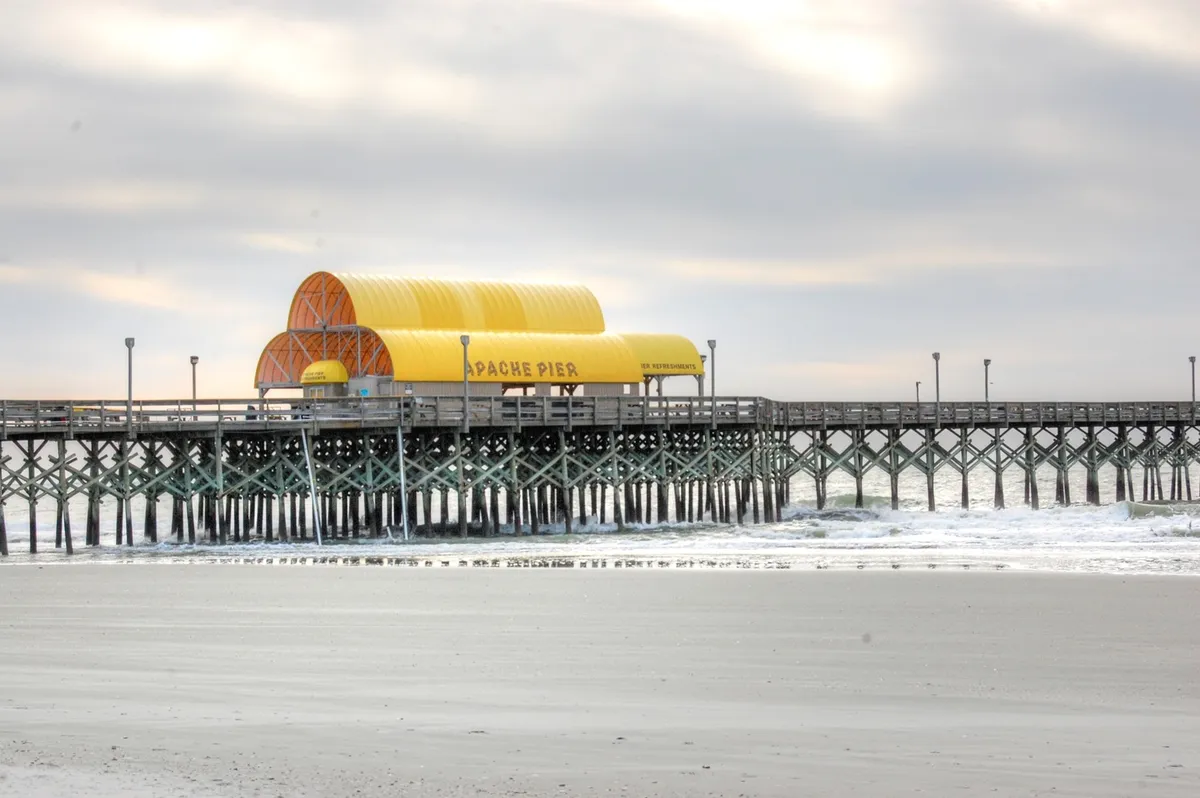 A scenic exterior shot highlighting the iconic Apache Pier with its striking yellow roof and sturdy wooden structure extending into the tranquil ocean. The setting sun casts a gentle glow, and the pier invites leisurely walks and moments of reflection along the water.