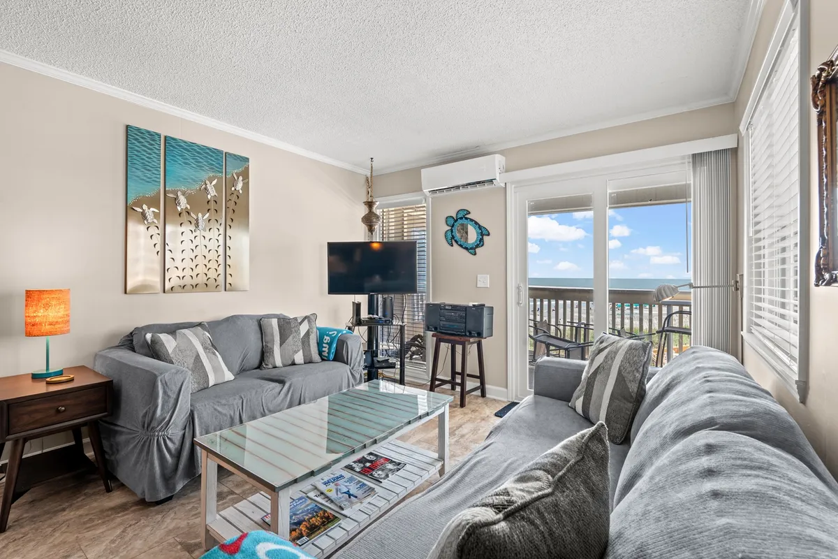 This inviting living room features a plush sofa and coffee table, with tasteful artwork on the wall. Sunlight pours through expansive windows, illuminating the cozy seating area, while a glimpse of the balcony and sunset view creates a relaxed, welcoming ambiance ideal for unwinding or socializing.