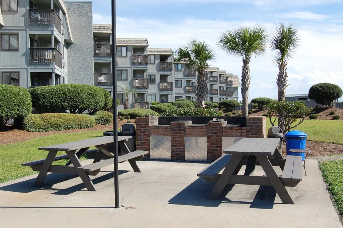 A welcoming outdoor grilling area features picnic tables and a built-in grill, shaded by palm trees and surrounded by lush landscaping. The communal setup encourages barbecues, picnics, and social gatherings in a relaxed, sunny environment.