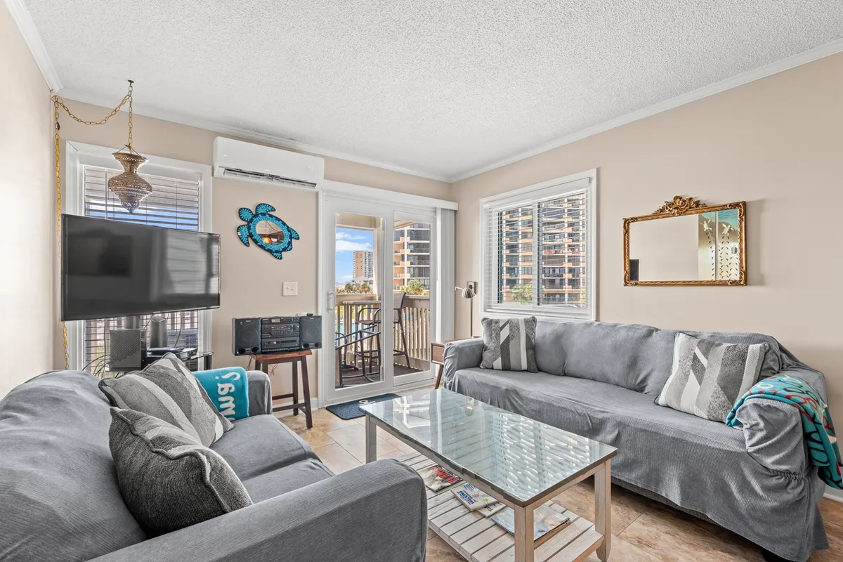 A spacious living room with a comfortable sofa, coffee table, television, decorative wall art, and large balcony doors letting in natural light. The room exudes a casual, welcoming vibe, ideal for relaxing or socializing after a day out.