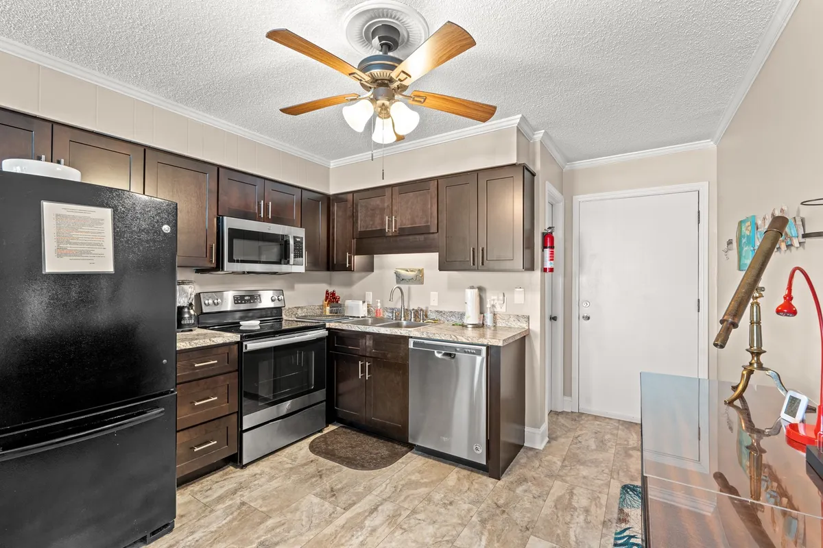 A modern kitchen equipped with a black refrigerator, stainless steel appliances, granite countertops, and a ceiling fan. The room is bright and practical, providing everything needed for preparing meals, enjoying coffee, or entertaining guests.