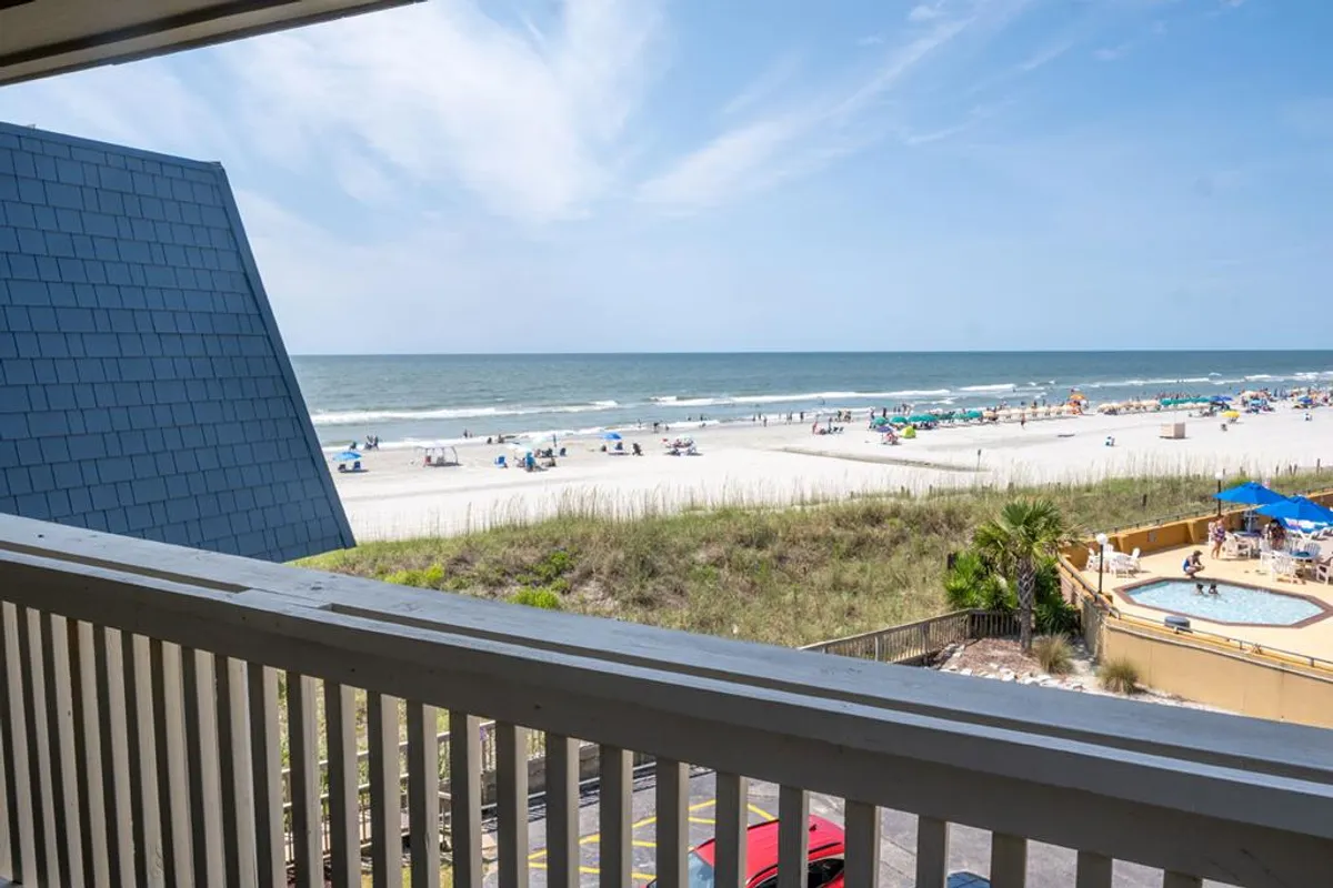 The image showcases a vibrant Myrtle Beach scene viewed from a condo balcony, featuring endless ocean waves, golden sand dunes, families enjoying the beach, and guests lounging by a sparkling swimming pool. The sun casts a warm glow, evoking the perfect blend of relaxation and fun by the coast.